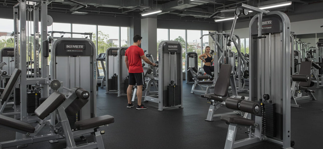 um homem de camisa vermelha fazwendo exercicio dentro de uma acade mia e uma mulher fazendo exercicio