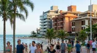 Pessoas caminhando pela orla de Canasvieiras, com prédios coloridos e palmeiras ao fundo.