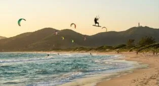 Kitesurfista salta sobre as ondas em praia com montanhas ao fundo e outros praticantes ao longe.