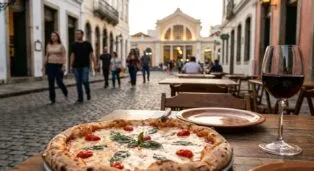 Pizza com manjericão e tomates em mesa de madeira, com pessoas caminhando em rua de paralelepípedos ao fundo.