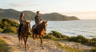 Casal montado em cavalos, passeando pela praia ao pôr do sol, com montanhas ao fundo.