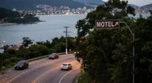 Placa de sinalização indicando direção para motel, vista de estrada com carros e paisagem de Florianópolis ao fundo.