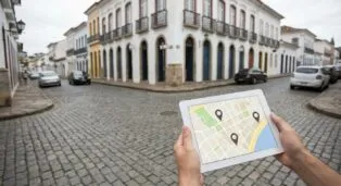 Pessoa segurando um tablet com mapa, em rua de paralelepípedo e casas coloridas ao fundo.