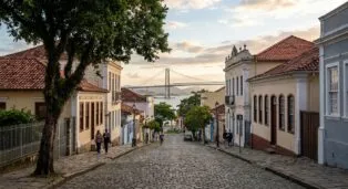 Rua de paralelepípedos com casas coloridas, árvores e a ponte ao fundo sob um céu com nuvens.