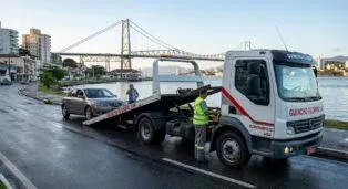 Guincho rebocando carro em Florianópolis, com ponte ao fundo e operador ao lado do veículo.