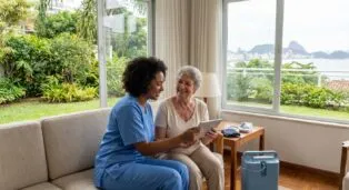 Profissional de saúde e idosa interagindo em sala iluminada, com vista para o exterior e equipamentos médicos ao fundo.