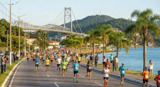 Corredores em diferentes camisetas coloridas participam da maratona à beira-mar, com a ponte ao fundo.