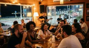Grupo de amigos sorrindo e se divertindo em um bar, enquanto músico toca violão ao fundo.