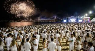 Multidão vestida de branco na praia, com fogos de artifício e palco ao fundo durante a virada do ano.