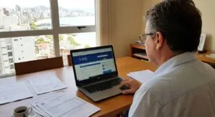 Homem sentado em mesa com laptop, consultando site sobre inscrição imobiliária, vista da cidade ao fundo.