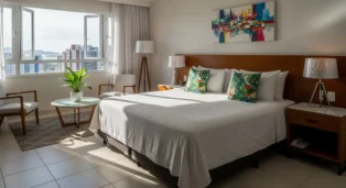 Quarto de hotel com cama de casal, decoração moderna e janelas amplas, vista para a cidade e plantas decorativas.
