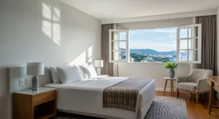 Quarto de hotel com cama grande, janelas abertas e vista para a cidade, decoração moderna e aconchegante.