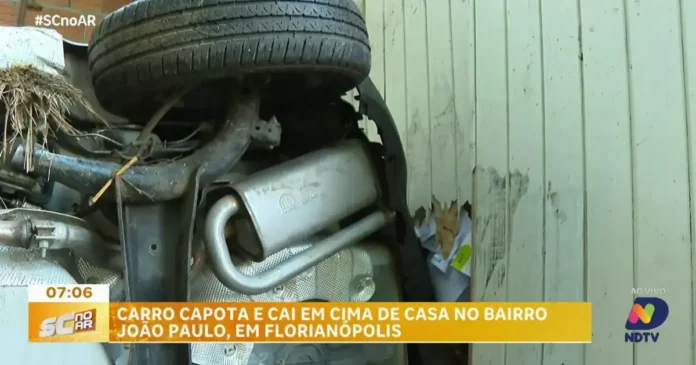Carro capotado em cima de uma casa, mostrando o teto do veículo e uma parede danificada.