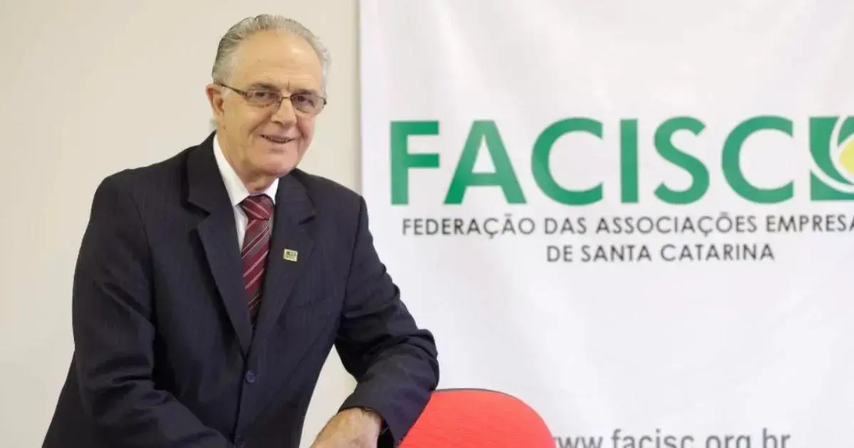 Um homem sorridente em um terno escuro, posicionado ao lado de um banner da FACISC com seu logo e texto.