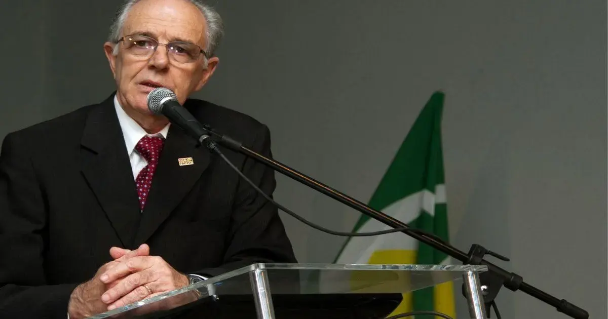 Alaor Francisco Tissot morre aos 87 anos em Florianópolis Homem de terno escuro falando ao microfone, com bandeira ao fundo e mãos sobre uma mesa de vidro.