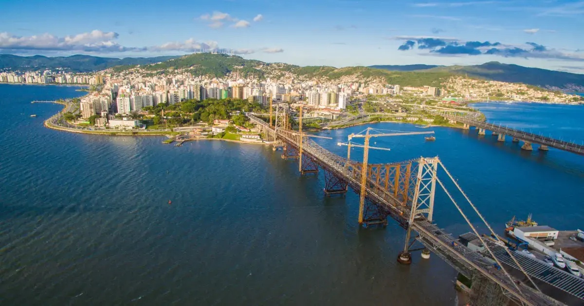 Vista aérea da cidade com prédios à beira de um rio e uma ponte de ferro em construção.