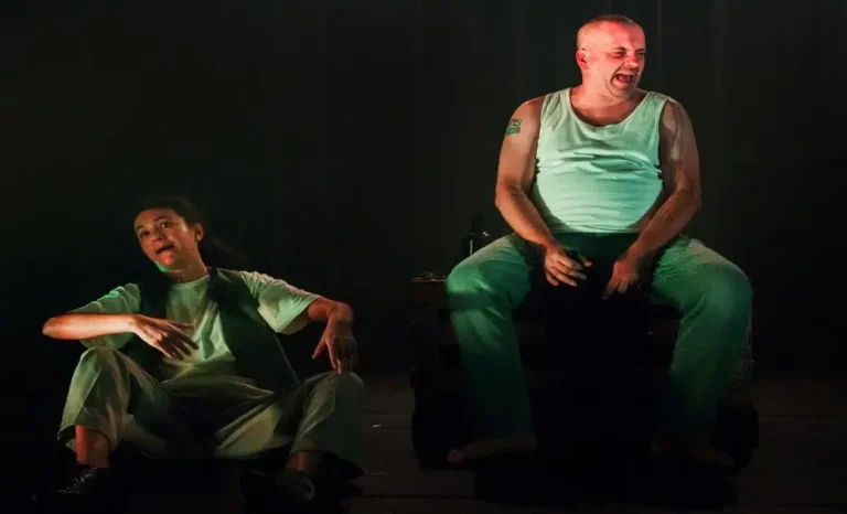 Duas pessoas em cena, uma sentada e outra em pé, aparentemente em um ambiente teatral.