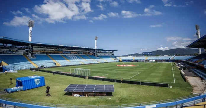 Estádio com campo de futebol, arquibancadas azuis e um céu claro. Painéis solares visíveis no primeiro plano.