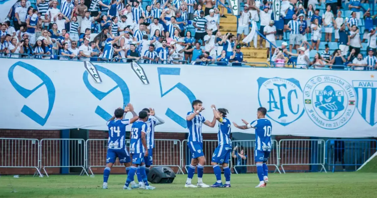 Avaí e Juventude: Preparativos para Confronto na Série B Torcedores comemoram na arquibancada enquanto jogadores do time celebram em campo, com bandeiras e camisas.