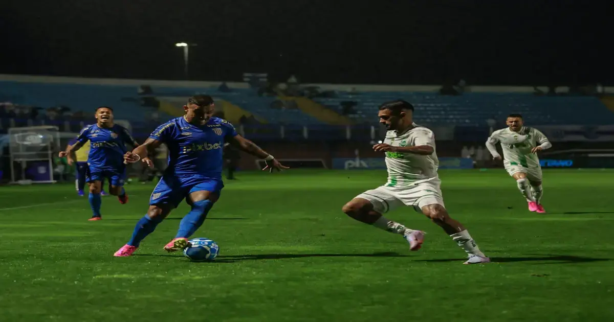 Jogadores em campo durante uma partida de futebol à noite, com um atleta em azul driblando um adversário em branco.