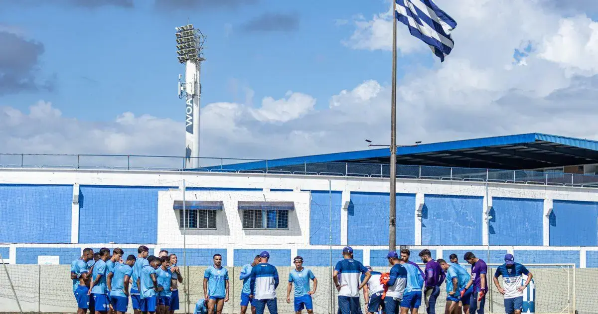 Treinamento de equipe de futebol em campo, com jogadores se reunindo próximo a uma bandeira e iluminação.