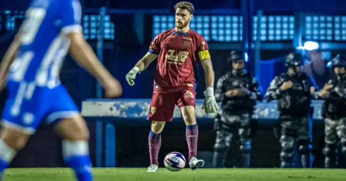 Goleiro em ação com uniformes marrom e vermelho, observando jogador adversário e segurança ao fundo.