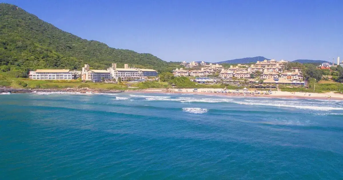 Praia com ondas suaves e areia, cercada por vegetação e edificações à beira-mar.