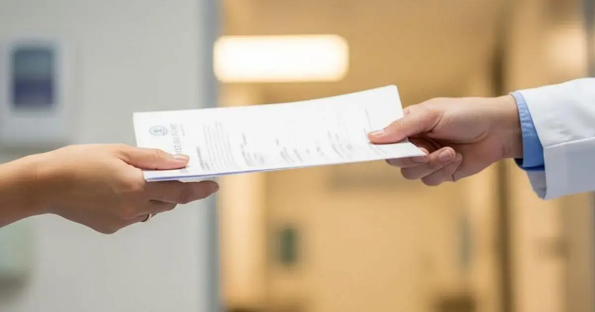 Boletim de Saúde Diário nas Unidades de Florianópolis Mão recebendo um documento de um profissional de saúde em um ambiente hospitalar.