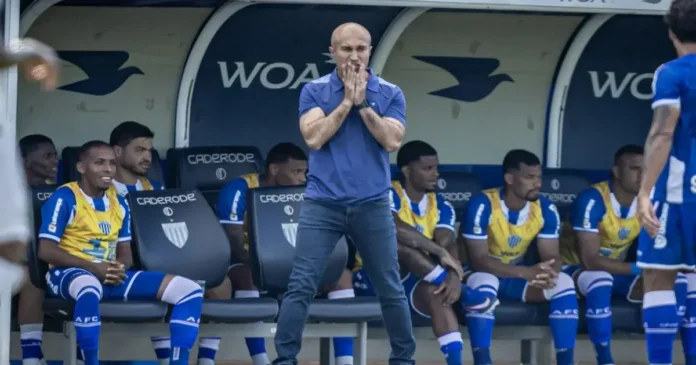 Treinador com expressão concentrada em campo, enquanto jogadores sentados no banco de reservas observam a partida.