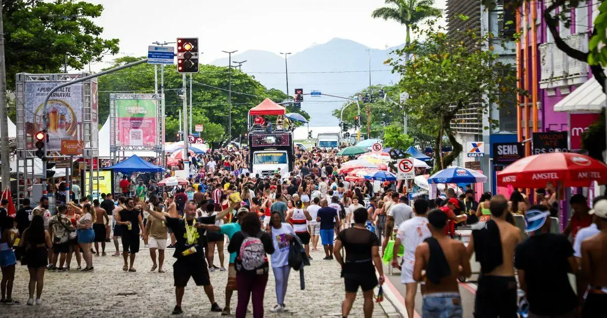 Rua lotada durante o Carnaval, com pessoas se divertindo, barracas e um trio elétrico ao fundo.