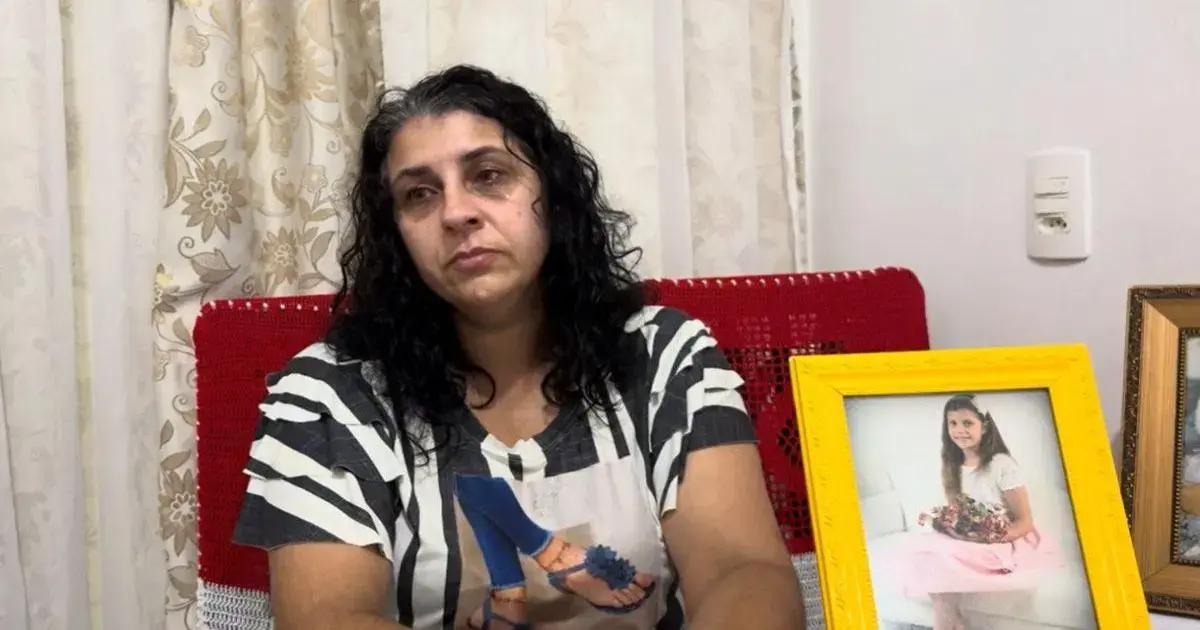 Mulher com expressão de tristeza sentada, ao lado de duas fotos emolduradas de uma menina, uma delas em um vestido rosa.