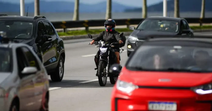 Motociclista trafegando entre veículos em uma rodovia, com montanhas ao fundo.