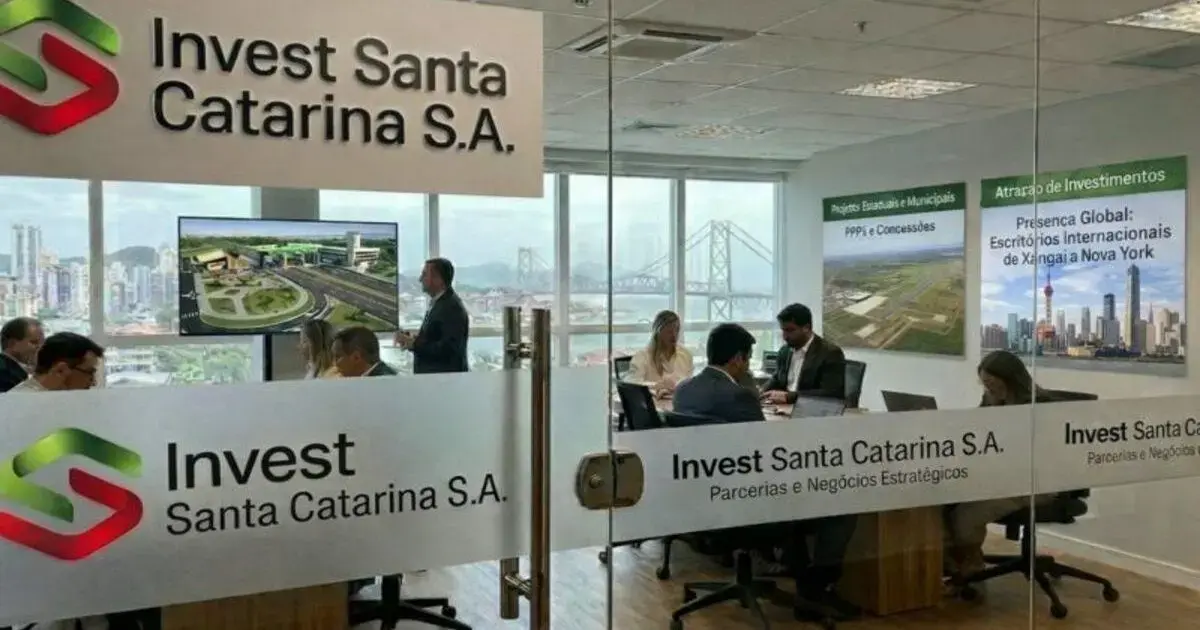 Reunião em escritório da Invest Santa Catarina S.A., com painel digital e vistas da cidade ao fundo.