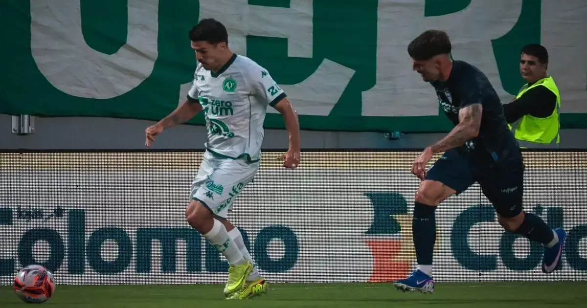 Jogadores em campo durante uma partida de futebol, com bandeira ao fundo e torcida visível.