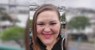 Mulher com cabelo longo e liso, usando blusa preta, sorrindo em um local externo com uma ponte e cidade ao fundo.