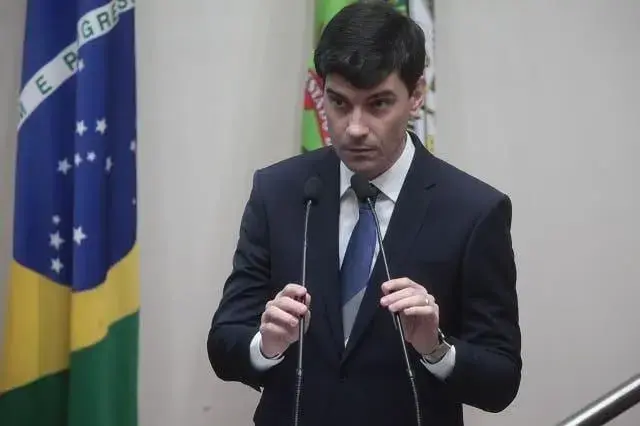 Homem em pé, com traje formal, falando ao microfone em ambiente legislativo; bandeiras ao fundo.