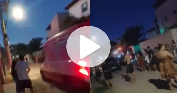 Grupo de pessoas em uma rua à noite, com uma ambulância ao fundo e iluminação pública visível.