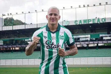 Jogador em campo de futebol, vestindo camisa listrada em verde e branco, fazendo gesto de vitória.