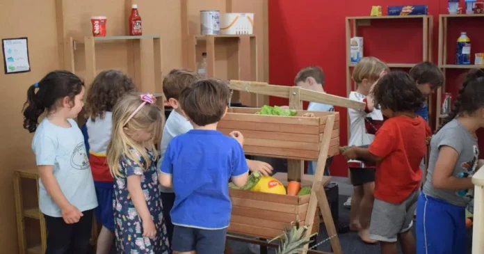 Crianças interagindo em um ambiente de aprendizagem, brincando com frutas e verduras em prateleiras de madeira.
