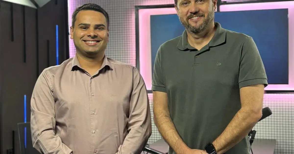 Dois homens sorrindo em um estúdio com fundo iluminado em tons de rosa e azul.