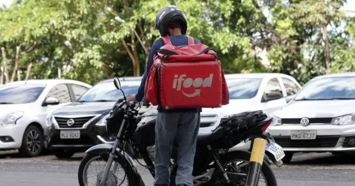 Entrega de refeição sendo realizada por motociclista com mochila vermelha do iFood em meio a carros estacionados.