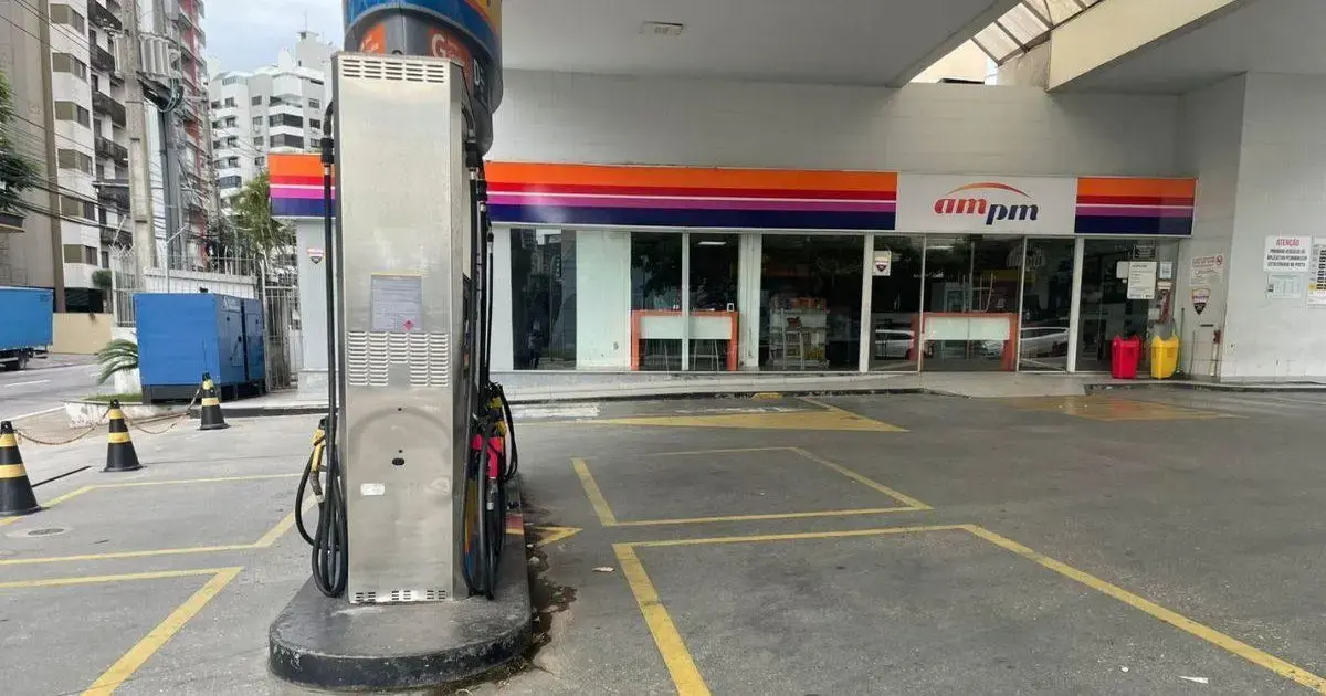 Posto de gasolina fechado com bomba de combustível visível e loja ampm ao fundo. Barreira de segurança e veículos estacionados.