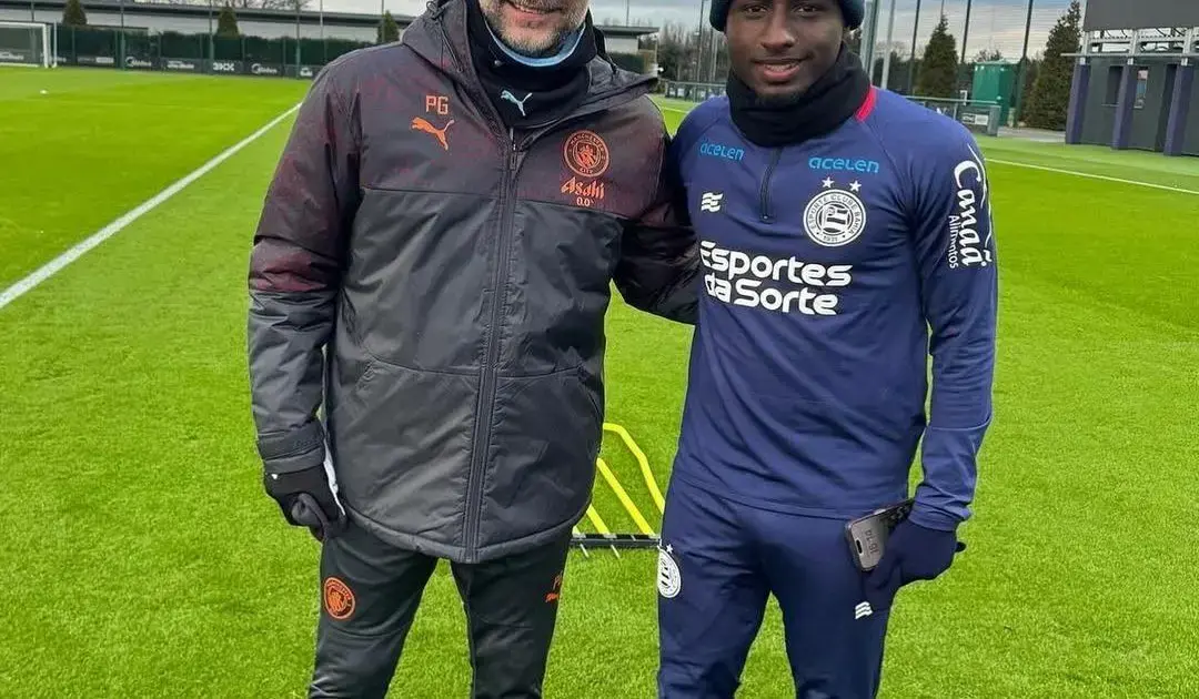 Figueirense anuncia contratação do lateral-esquerdo Ryan Carlos Dois homens sorrindo em um campo de futebol, um vestindo jaqueta preta e o outro em uniforme azul.