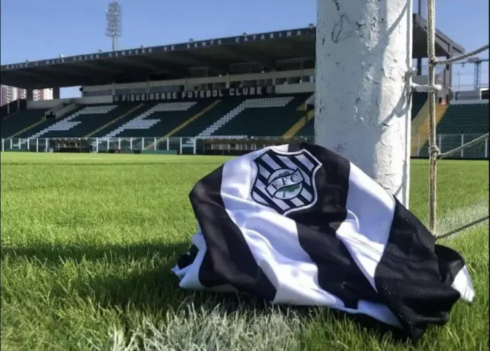 Camisa do clube de futebol em campo, com o estádio ao fundo e a rede do gol visível.