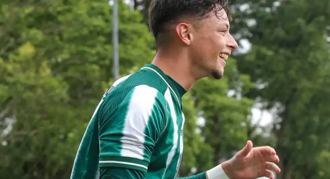 Jogador sorridente, vestindo camisa verde com detalhes brancos, em campo cercado por vegetação.