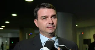 Pessoas conversando em ambiente interno, um homem em destaque falando ao microfone e cercado por aparelhos de gravação.