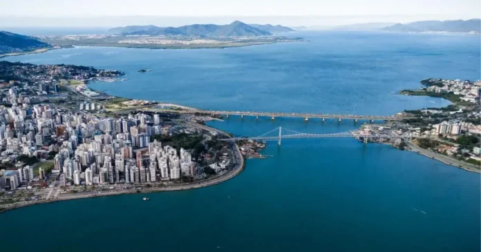 Vista aérea de uma cidade costeira com prédios altos, praia, um rio e montanhas ao fundo, além de uma ponte sobre a água.