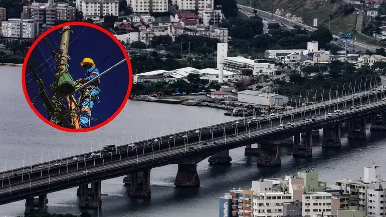 Trabalho em altura em fiação elétrica, com a ponte ao fundo e edifícios residenciais nas margens do rio.