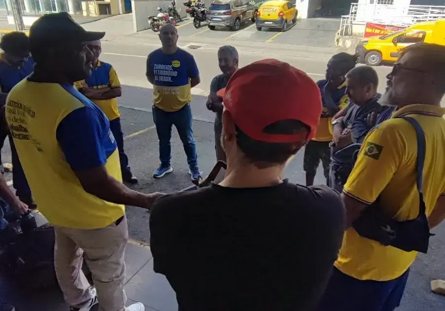 Grupo de trabalhadores em reunião, usando camisetas azuis e amarelas, em frente a um prédio.
