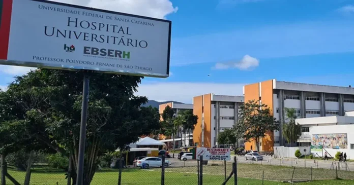 Placa do Hospital Universitário com o nome, localização e informações sobre a instituição; edifícios ao fundo.